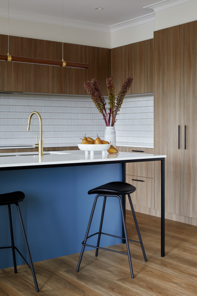 Photo of a small midcentury l-shaped open plan kitchen in Melbourne with an undermount sink, flat-panel cabinets, medium wood cabinets, quartz benchtops, white splashback, mosaic tile splashback, black appliances, laminate floors, with island, brown floor and white benchtop.