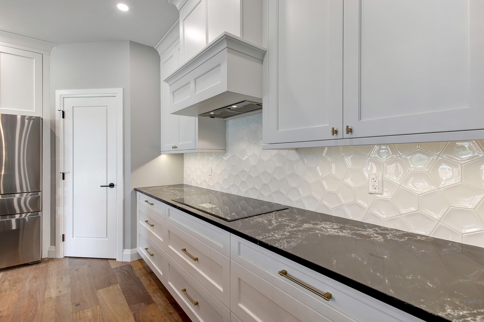 Example of a mid-sized transitional l-shaped medium tone wood floor and brown floor open concept kitchen design in Edmonton with an undermount sink, recessed-panel cabinets, white cabinets, quartz countertops, white backsplash, ceramic backsplash, stainless steel appliances, an island and black countertops