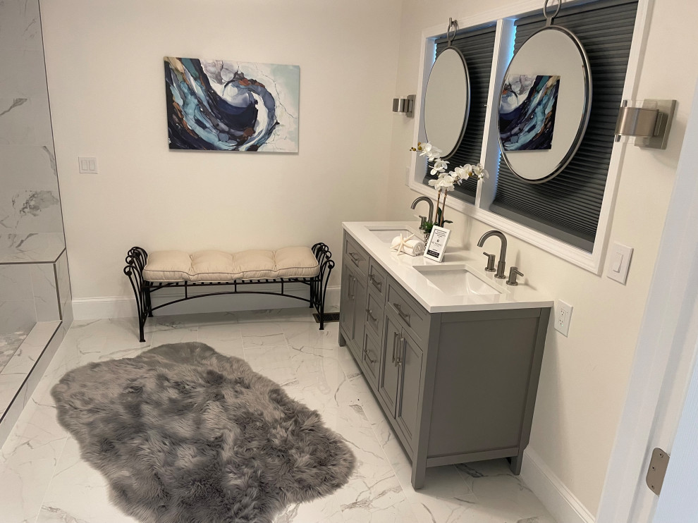 Modern bathroom with a gray vanity, round mirrors, a cushioned bench, marble-style tile, and a gray rug.
