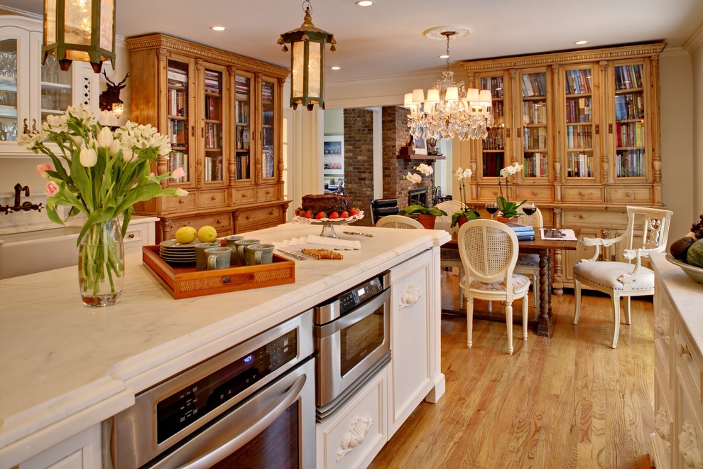 Photo of an eat-in kitchen in New York with glass-front cabinets, stainless steel appliances, a farmhouse sink, white cabinets and marble benchtops.