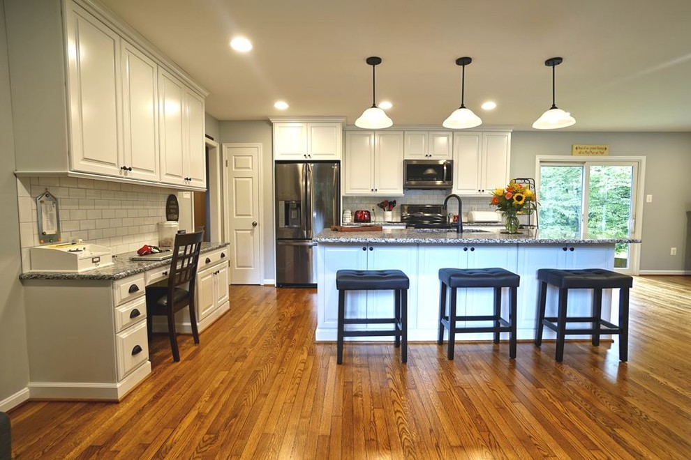 Photo of an expansive country eat-in kitchen in DC Metro with an undermount sink, white cabinets, granite benchtops, white splashback, subway tile splashback, stainless steel appliances, medium hardwood floors, with island, brown floor and black benchtop.