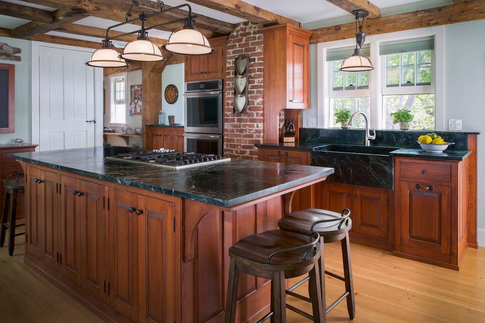 This is an example of a large country kitchen in Portland Maine with soapstone benchtops, with island, a farmhouse sink, raised-panel cabinets, medium wood cabinets, stainless steel appliances, light hardwood floors and beige floor.
