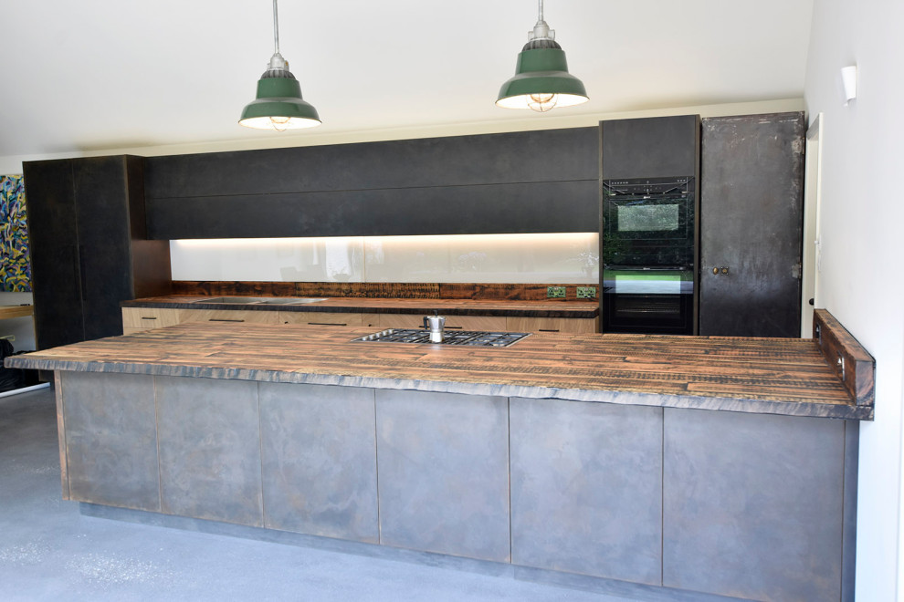 Inspiration for a large industrial single-wall open plan kitchen in Oxfordshire with a double-bowl sink, flat-panel cabinets, light wood cabinets, wood benchtops, white splashback, glass sheet splashback, black appliances, concrete floors, with island, grey floor and multi-coloured benchtop.
