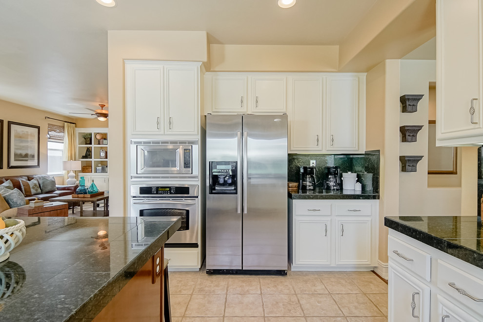 Photo of a mid-sized traditional l-shaped eat-in kitchen in San Diego with a double-bowl sink, recessed-panel cabinets, white cabinets, granite benchtops, black splashback, stone tile splashback, stainless steel appliances, ceramic floors, with island and beige floor.