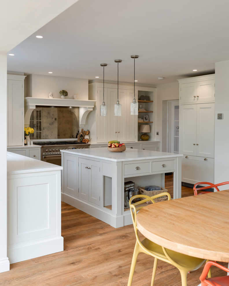 Large farmhouse grey and white l-shaped open plan kitchen in Hertfordshire with an integrated sink, shaker cabinets, white cabinets, quartz worktops, white splashback, stainless steel appliances, light hardwood flooring, an island, brown floors, white worktops and a chimney breast.