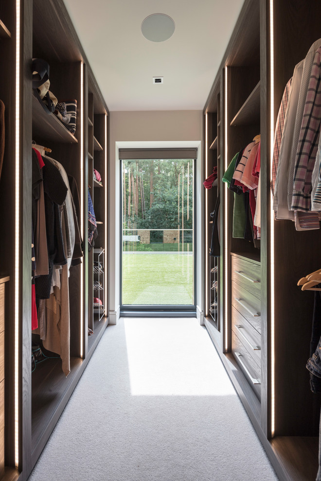 Photo of a contemporary gender-neutral walk-in wardrobe in Surrey with flat-panel cabinets, dark wood cabinets, carpet and white floor.