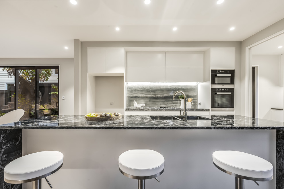 Mid-sized contemporary single-wall eat-in kitchen in Auckland with an undermount sink, flat-panel cabinets, white cabinets, marble benchtops, black splashback, marble splashback, stainless steel appliances, light hardwood floors, with island and black benchtop.