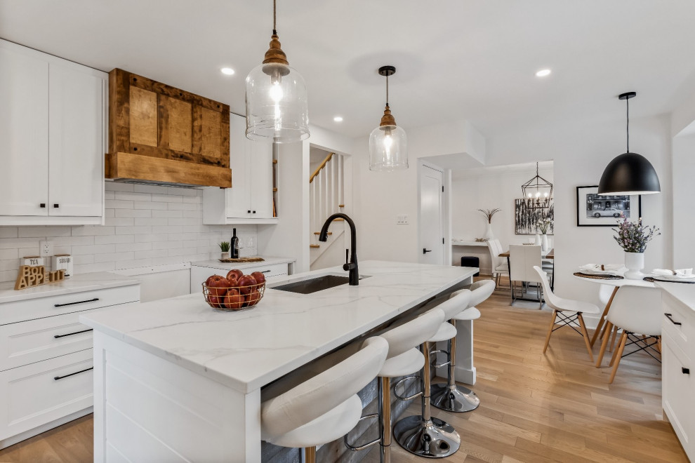 Diseño de cocina campestre, grande, cerrada, con fregadero bajoencimera, armarios estilo shaker, puertas de armario blancas, encimera de cuarzo compacto, salpicadero blanco, salpicadero de azulejos tipo metro, suelo de madera clara, una isla, suelo marrón y encimeras blancas