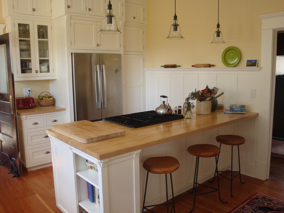 Photo of a small arts and crafts galley separate kitchen in Seattle with a farmhouse sink, shaker cabinets, white cabinets, wood benchtops, white splashback, ceramic splashback, stainless steel appliances, medium hardwood floors, a peninsula, brown floor and beige benchtop.