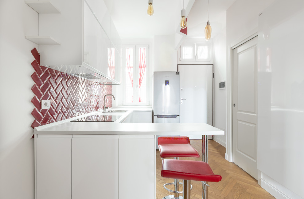 Design ideas for a mid-sized contemporary u-shaped open plan kitchen in Milan with a drop-in sink, flat-panel cabinets, white cabinets, red splashback, subway tile splashback, stainless steel appliances, light hardwood floors, a peninsula, brown floor and white benchtop.
