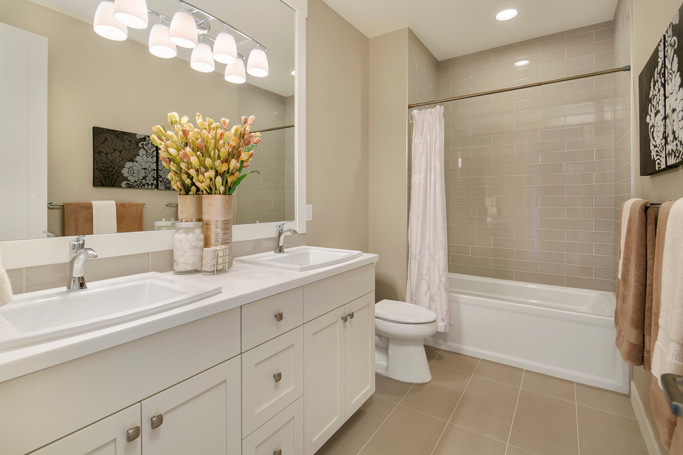 This is an example of an arts and crafts bathroom in Seattle with white cabinets, an alcove tub, a shower/bathtub combo, a two-piece toilet, gray tile, ceramic tile, beige walls, porcelain floors, a drop-in sink, solid surface benchtops and recessed-panel cabinets.