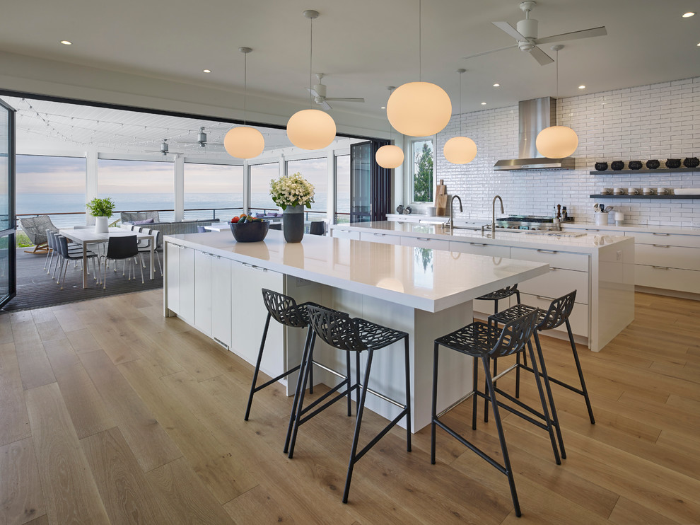 Photo of a large contemporary galley open plan kitchen in Other with an undermount sink, flat-panel cabinets, white cabinets, solid surface benchtops, white splashback, subway tile splashback, stainless steel appliances, light hardwood floors, multiple islands and yellow floor.