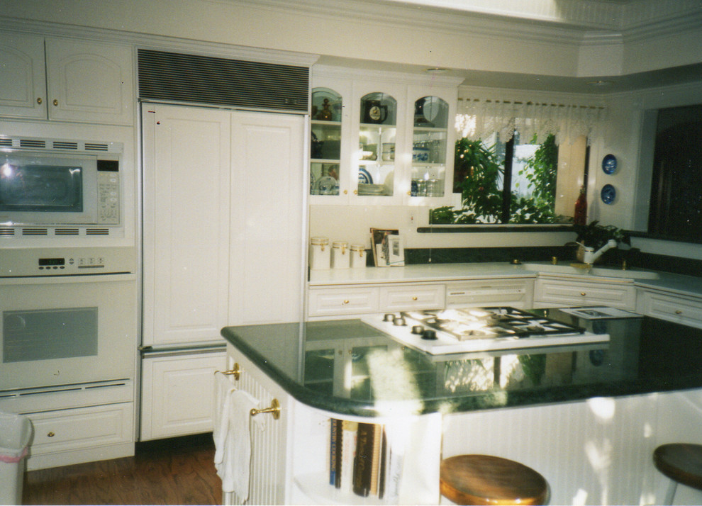 Inspiration for a transitional l-shaped medium tone wood floor enclosed kitchen remodel in Orange County with an integrated sink, raised-panel cabinets, white cabinets, solid surface countertops, white backsplash, ceramic backsplash, white appliances and an island