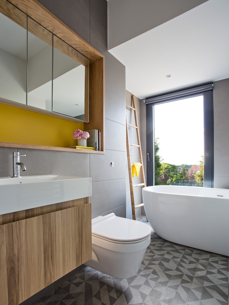 This is an example of a mid-sized contemporary bathroom in London with flat-panel cabinets, brown cabinets, a freestanding tub, a wall-mount toilet, gray tile, grey walls, ceramic floors and a wall-mount sink.