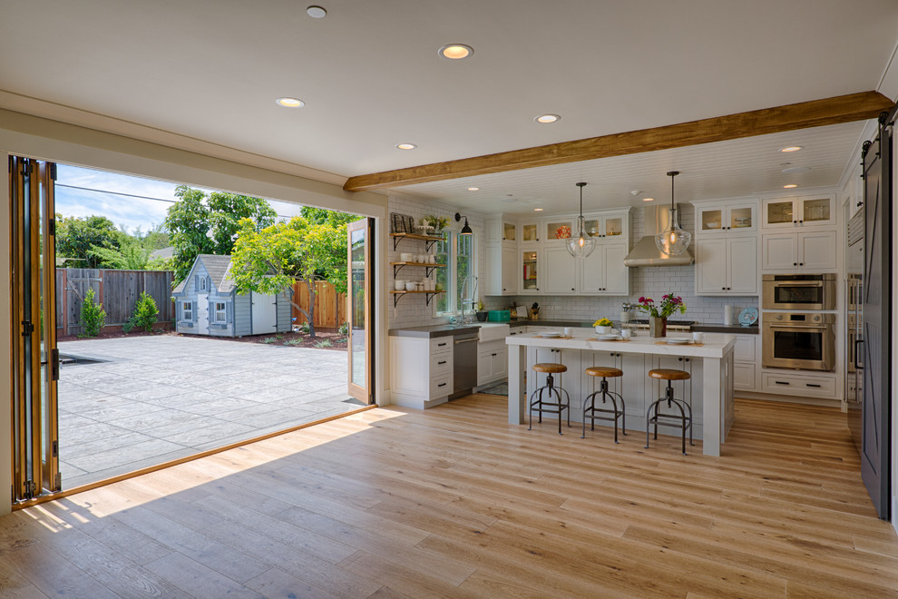 Mid-sized country u-shaped open plan kitchen in San Francisco with a farmhouse sink, white splashback, porcelain splashback, stainless steel appliances, with island, beige floor and light hardwood floors.