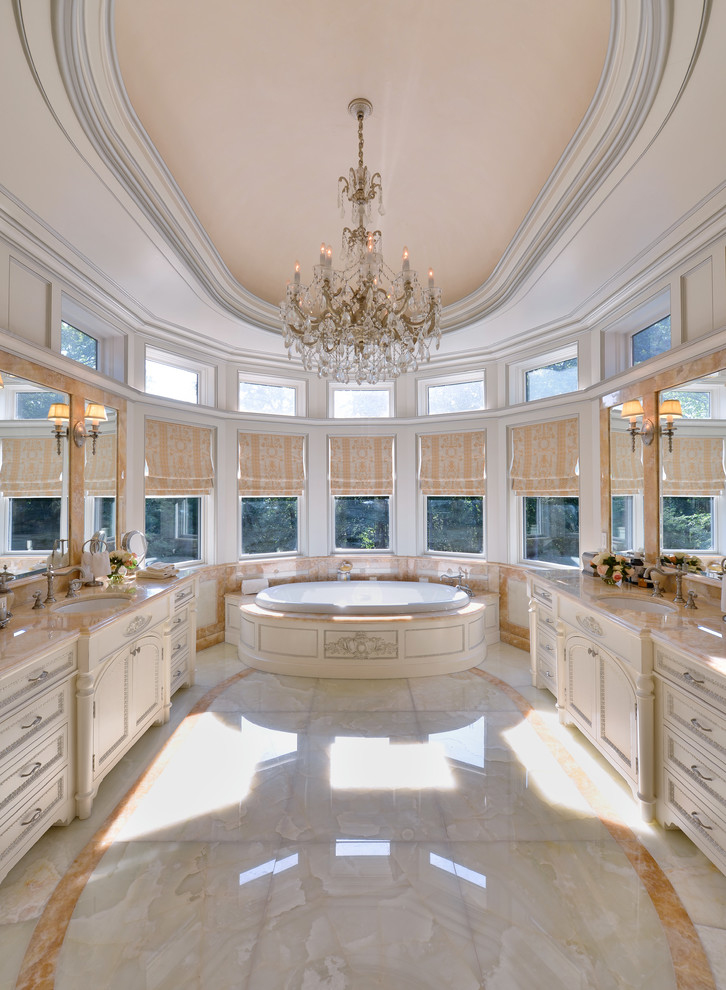 Photo of an expansive traditional master bathroom in New York with an undermount sink, furniture-like cabinets, light wood cabinets, onyx benchtops, a drop-in tub, a one-piece toilet, beige tile, stone slab, beige walls and marble floors.