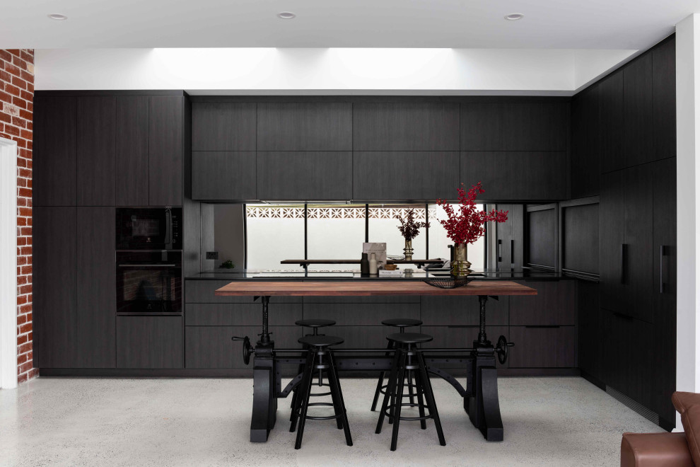 Photo of a mid-sized contemporary l-shaped eat-in kitchen in Perth with a drop-in sink, recessed-panel cabinets, black cabinets, quartz benchtops, black splashback, mirror splashback, black appliances, no island, black benchtop, concrete floors and grey floor.