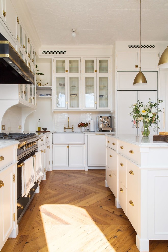 Design ideas for a modern l-shaped kitchen in Los Angeles with a farmhouse sink, shaker cabinets, white cabinets, white splashback, panelled appliances, medium hardwood floors, with island, brown floor and white benchtop.