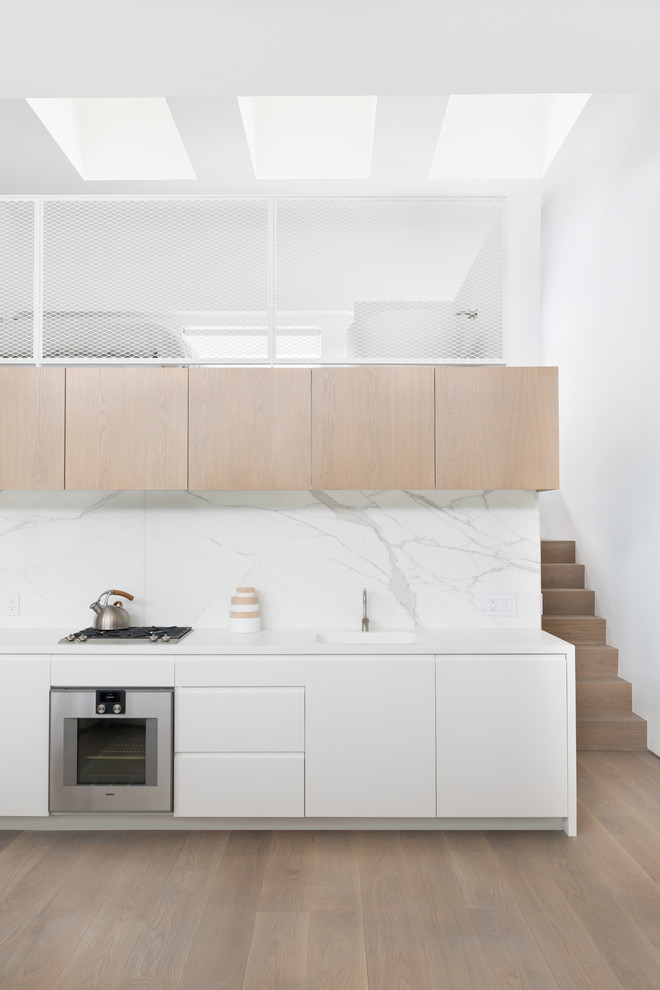 Design ideas for a scandinavian single-wall kitchen in New York with flat-panel cabinets, white cabinets, white splashback, stone slab splashback, stainless steel appliances, medium hardwood floors, no island, brown floor and white benchtop.