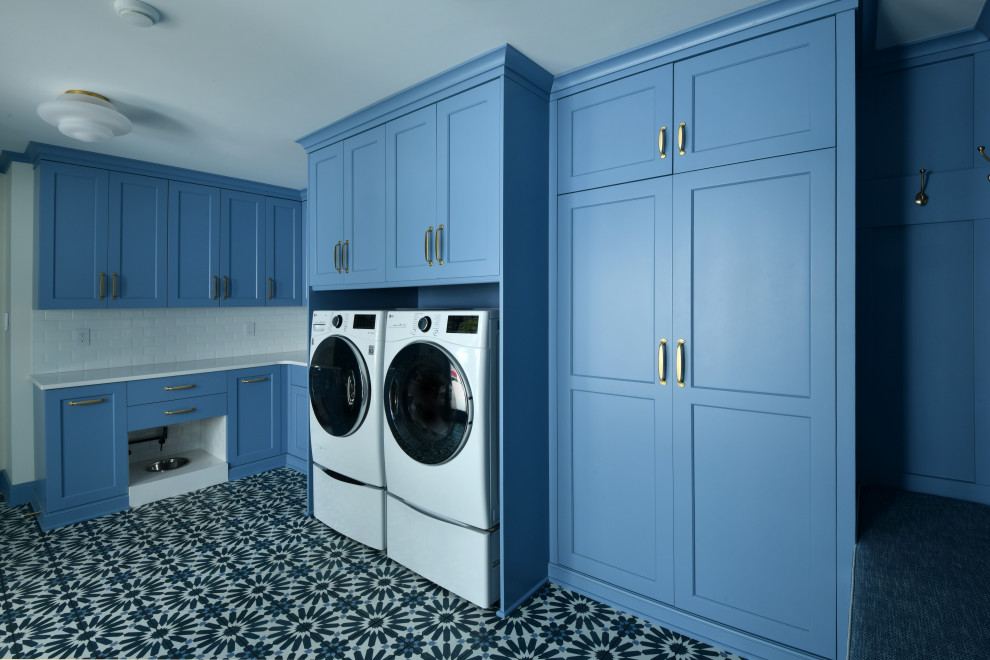 他の地域にある巨大なトランジショナルスタイルのおしゃれな家事室 (コの字型、アンダーカウンターシンク、フラットパネル扉のキャビネット、青いキャビネット、クオーツストーンカウンター、白いキッチンパネル、サブウェイタイルのキッチンパネル、白い壁、コンクリートの床、左右配置の洗濯機・乾燥機、青い床、白いキッチンカウンター) の写真
