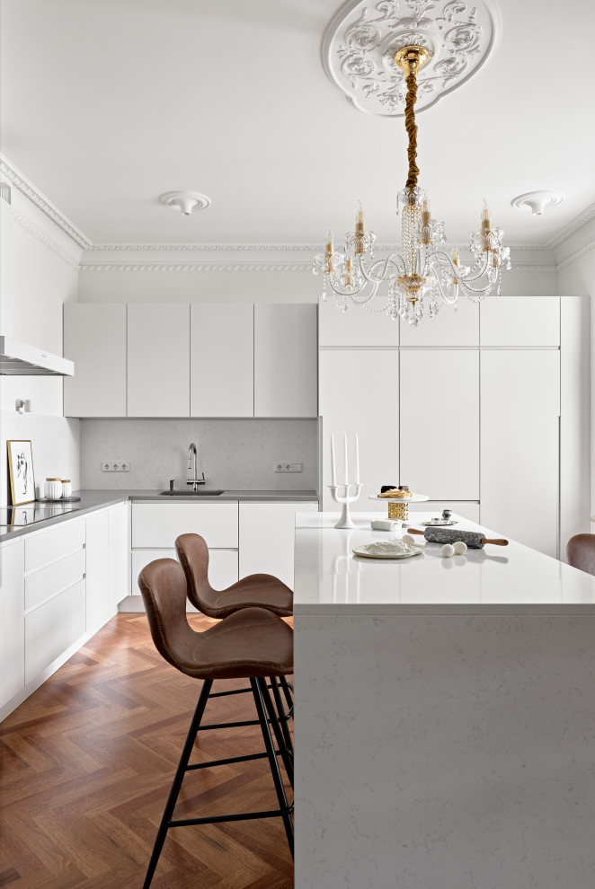 This is an example of a large transitional single-wall eat-in kitchen in Saint Petersburg with an undermount sink, flat-panel cabinets, white cabinets, solid surface benchtops, white splashback, engineered quartz splashback, white appliances, medium hardwood floors, with island, brown floor and grey benchtop.