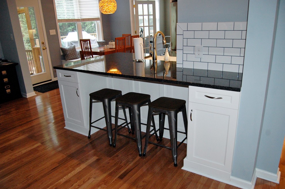 This is an example of a contemporary eat-in kitchen in Raleigh with a farmhouse sink, shaker cabinets, white cabinets, granite benchtops, white splashback, subway tile splashback, stainless steel appliances and medium hardwood floors.