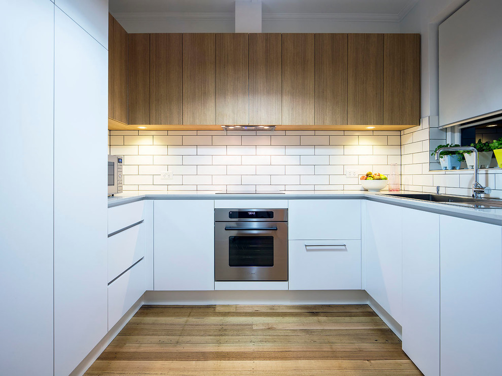 Inspiration for a small contemporary u-shaped separate kitchen in Melbourne with a drop-in sink, flat-panel cabinets, white cabinets, white splashback, subway tile splashback, panelled appliances, medium hardwood floors and no island.