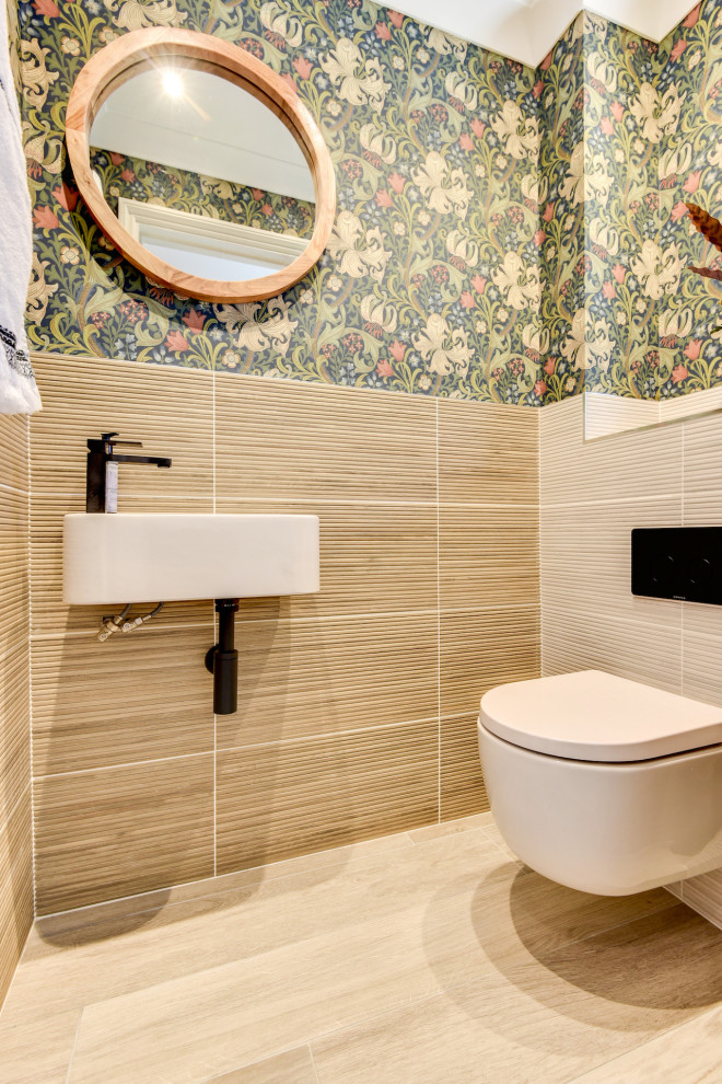 This is an example of a small modern powder room in Sussex with a one-piece toilet, beige tile, ceramic tile, multi-coloured walls, ceramic floors, a wall-mount sink, tile benchtops, beige floor, beige benchtops and wallpaper.