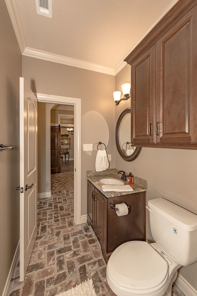 Photo of a mid-sized country 3/4 bathroom in Austin with shaker cabinets, medium wood cabinets, a two-piece toilet, brown tile, travertine, beige walls, brick floors, an undermount sink, granite benchtops and brown floor.