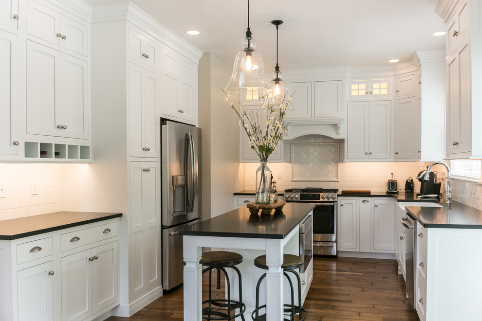 Photo of a traditional u-shaped eat-in kitchen in Philadelphia with a farmhouse sink, shaker cabinets, white cabinets, white splashback, subway tile splashback, stainless steel appliances, dark hardwood floors, with island, brown floor, black benchtop and soapstone benchtops.