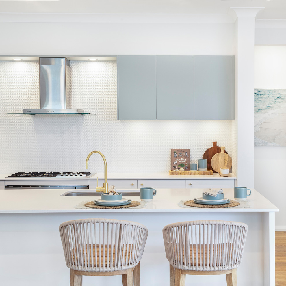 Beach style galley kitchen in Sydney with an undermount sink, flat-panel cabinets, grey cabinets, white splashback, mosaic tile splashback, stainless steel appliances, medium hardwood floors, with island, brown floor and white benchtop.