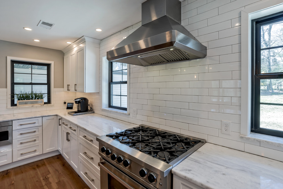 This is an example of a mid-sized transitional u-shaped eat-in kitchen in Philadelphia with a farmhouse sink, shaker cabinets, white cabinets, quartzite benchtops, white splashback, porcelain splashback, stainless steel appliances, light hardwood floors, with island, brown floor and white benchtop.