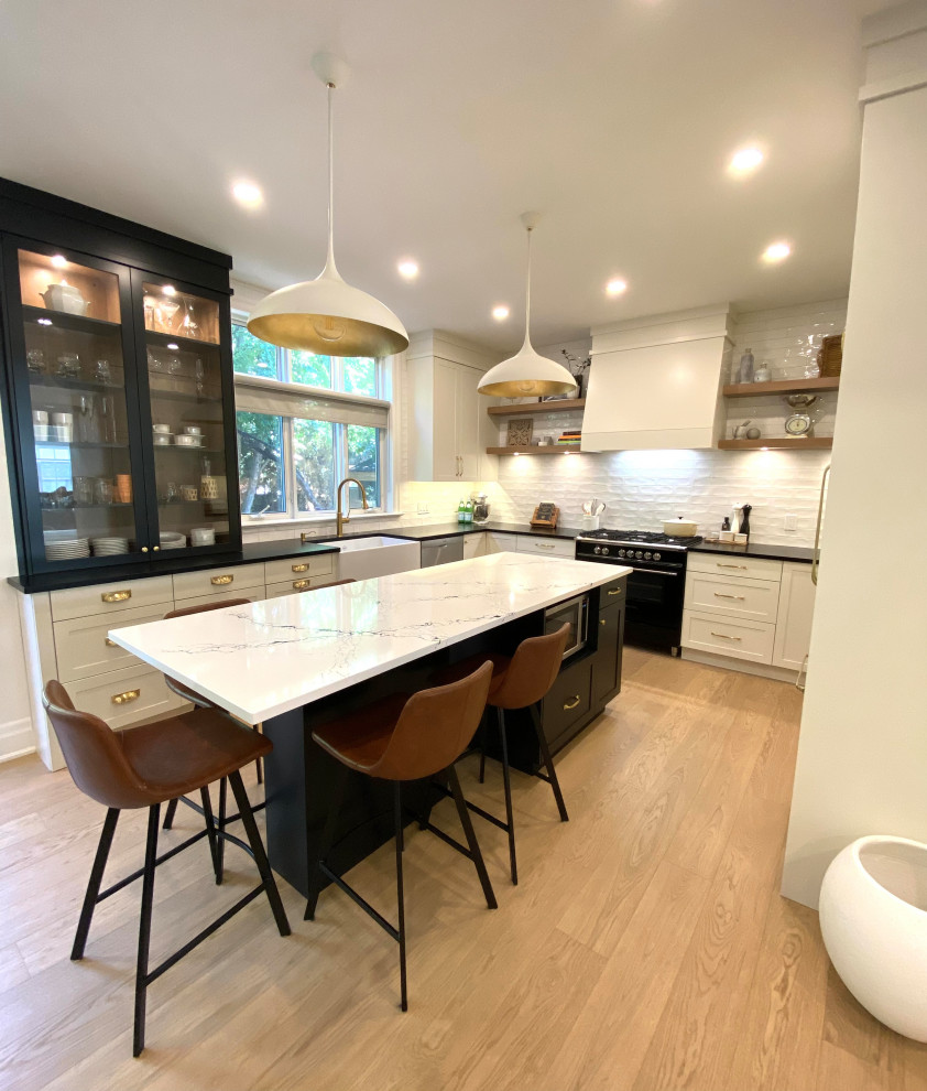 Example of a mid-sized transitional l-shaped light wood floor open concept kitchen design in Toronto with a farmhouse sink, shaker cabinets, white cabinets, quartz countertops, white backsplash, porcelain backsplash, an island and black countertops