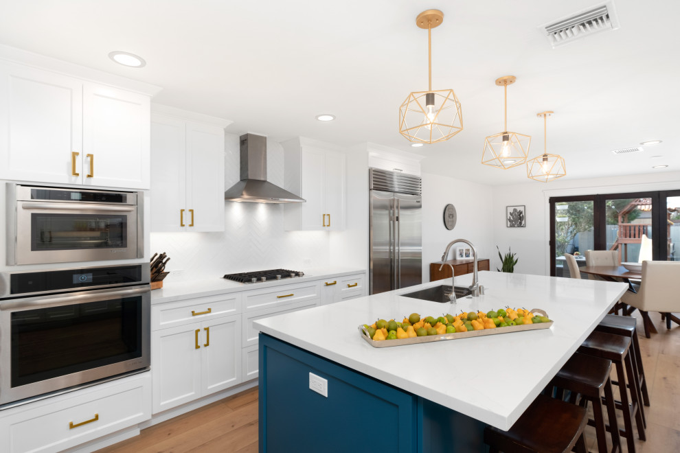 Expansive contemporary single-wall eat-in kitchen in San Diego with a drop-in sink, recessed-panel cabinets, white cabinets, quartzite benchtops, white splashback, ceramic splashback, stainless steel appliances, medium hardwood floors, with island, brown floor and white benchtop.