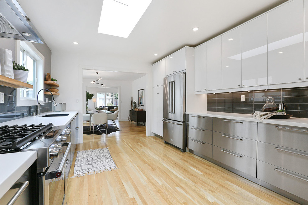 This is an example of a large modern galley separate kitchen in San Francisco with an undermount sink, flat-panel cabinets, white cabinets, black splashback, glass tile splashback, stainless steel appliances, light hardwood floors, no island, beige floor, quartz benchtops and white benchtop.