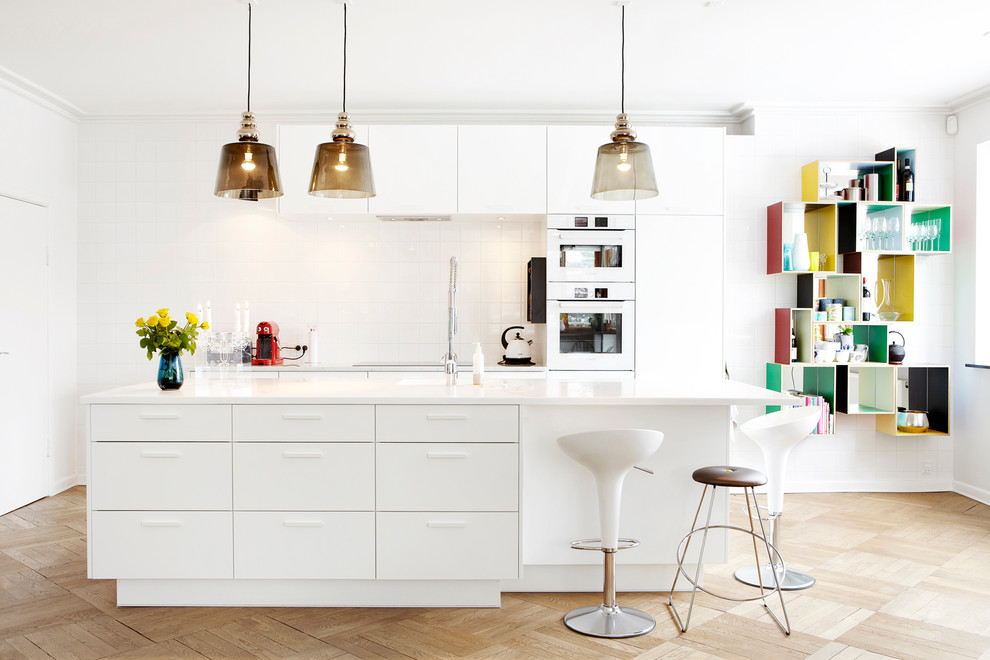 Large contemporary galley eat-in kitchen in Copenhagen with flat-panel cabinets, white cabinets, white splashback, white appliances, light hardwood floors, with island, a drop-in sink, recycled glass benchtops and porcelain splashback.