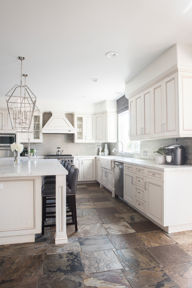Inspiration for a mid-sized contemporary l-shaped eat-in kitchen in Orange County with white cabinets, stainless steel appliances, with island, brown floor, a farmhouse sink, quartz benchtops, grey splashback, glass tile splashback, slate floors and recessed-panel cabinets.