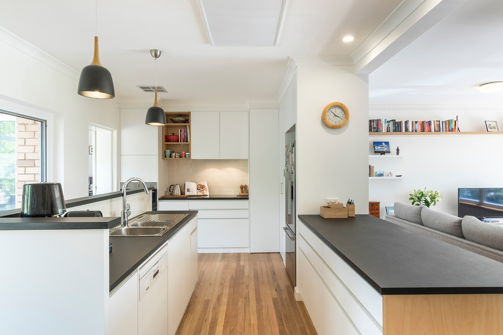 Design ideas for a mid-sized contemporary u-shaped open plan kitchen in Canberra - Queanbeyan with a double-bowl sink, flat-panel cabinets, white cabinets, beige splashback, ceramic splashback, stainless steel appliances, black benchtop, medium hardwood floors, a peninsula and brown floor.