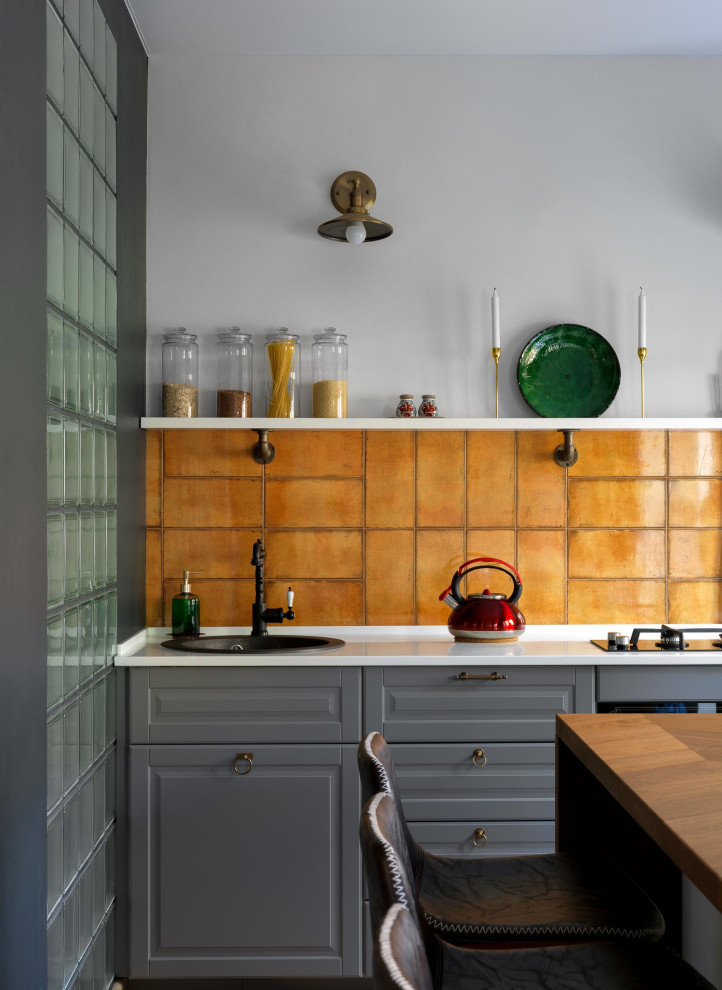 Photo of a small eclectic single-wall open plan kitchen in Moscow with a drop-in sink, recessed-panel cabinets, grey cabinets, solid surface benchtops, orange splashback, ceramic splashback, black appliances, medium hardwood floors, with island, multi-coloured floor and white benchtop.