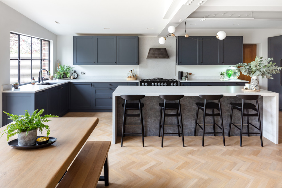 Photo of a mid-sized contemporary l-shaped open plan kitchen in London with a drop-in sink, recessed-panel cabinets, black cabinets, marble benchtops, white splashback, stone tile splashback, black appliances, light hardwood floors, with island, brown floor, white benchtop and exposed beam.