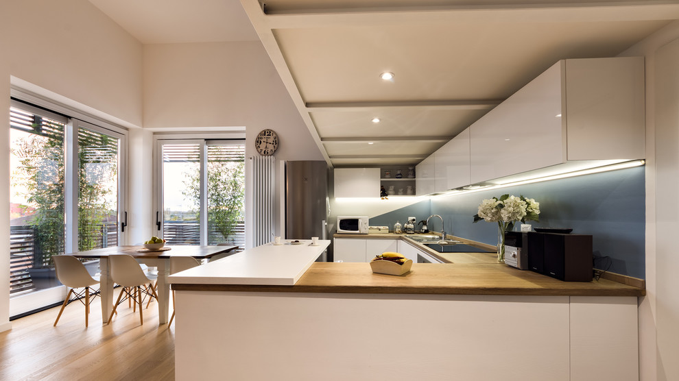 Photo of a large contemporary u-shaped eat-in kitchen in Milan with a drop-in sink, flat-panel cabinets, white cabinets, wood benchtops, blue splashback, stainless steel appliances, glass sheet splashback, medium hardwood floors, a peninsula and beige floor.