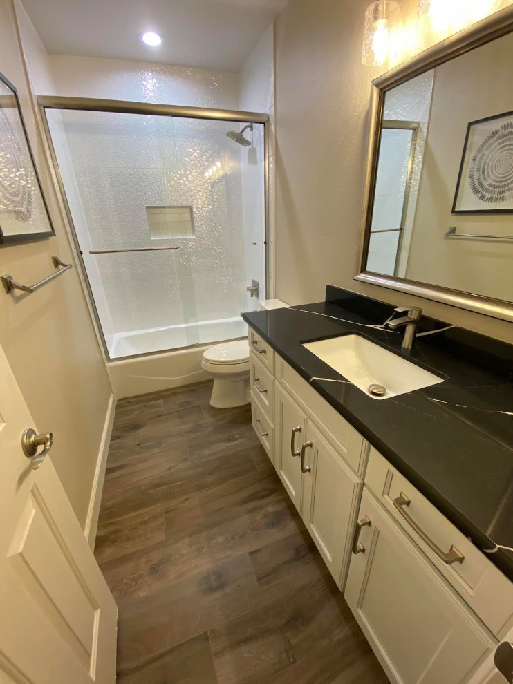 This is an example of a mid-sized modern bathroom in Phoenix with recessed-panel cabinets, white cabinets, an alcove tub, a shower/bathtub combo, a one-piece toilet, white tile, porcelain tile, beige walls, wood-look tile, an undermount sink, engineered quartz benchtops, multi-coloured floor, a sliding shower screen, black benchtops, a single vanity and a built-in vanity.