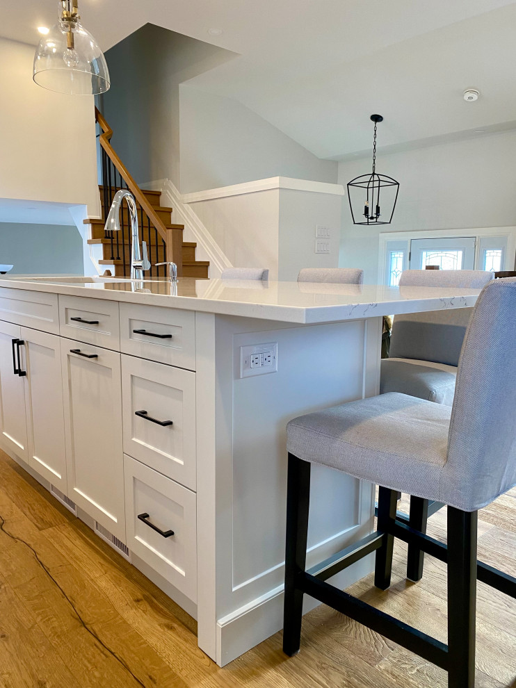 Mid-sized country l-shaped open plan kitchen in Toronto with an undermount sink, shaker cabinets, white cabinets, quartz benchtops, white splashback, engineered quartz splashback, stainless steel appliances, medium hardwood floors, with island, white benchtop and vaulted.