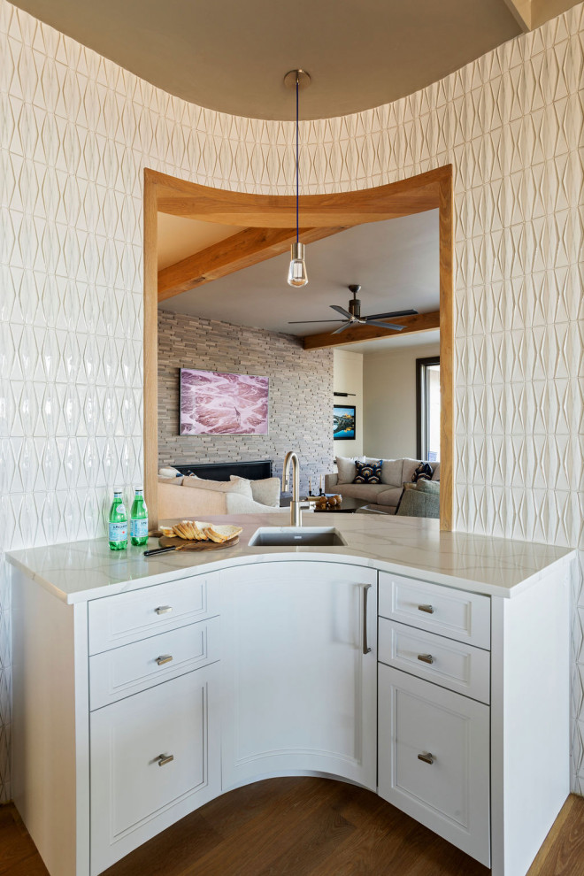 Photo of a small beach style u-shaped wet bar in Charleston with a built-in sink, white cabinets, marble worktops, white splashback, porcelain splashback, light hardwood flooring, brown floors, white worktops and a feature wall.