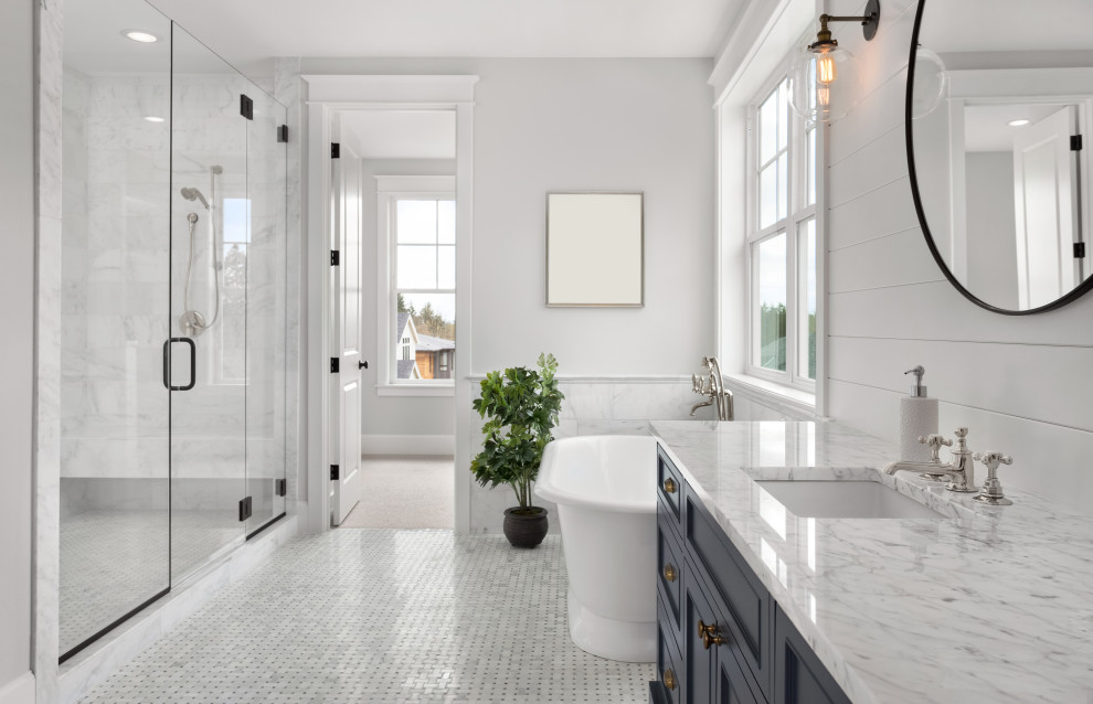 Идея дизайна: большая главная ванная комната: освещение в стиле неоклассика (современная классика) с фасадами в стиле шейкер, синими фасадами, отдельно стоящей ванной, угловым душем, раздельным унитазом, мраморной плиткой, белыми стенами, полом из мозаичной плитки, врезной раковиной, мраморной столешницей, серым полом, душем с распашными дверями, серой столешницей, тумбой под две раковины, встроенной тумбой, сиденьем для душа и окном
