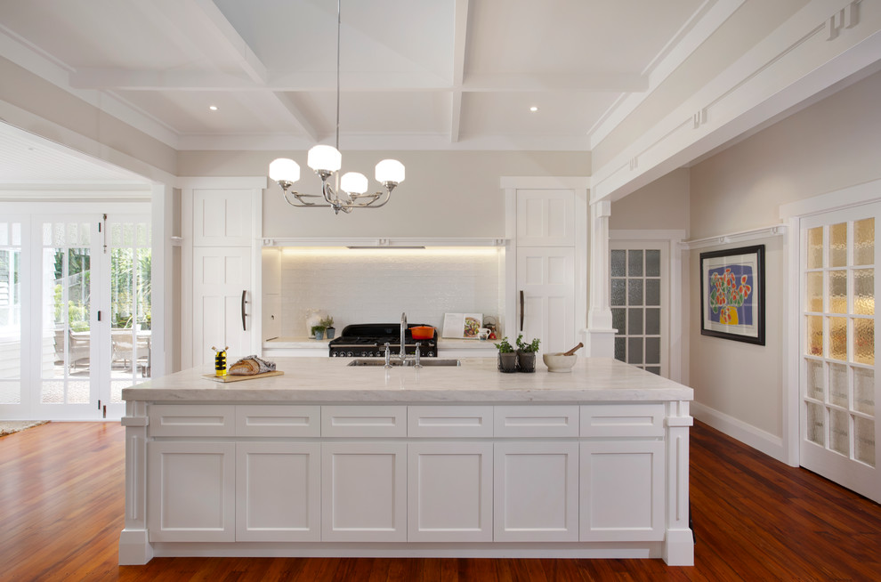 This is an example of a traditional galley open plan kitchen in Auckland with white cabinets, white splashback, subway tile splashback, black appliances, medium hardwood floors, with island, an undermount sink, recessed-panel cabinets and solid surface benchtops.