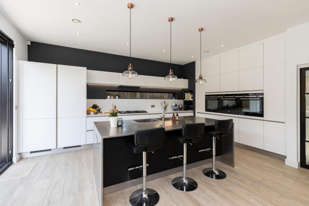 This is an example of a large contemporary l-shaped open plan kitchen in Hertfordshire with flat-panel cabinets, white cabinets, stainless steel worktops, black appliances, an island and feature lighting.