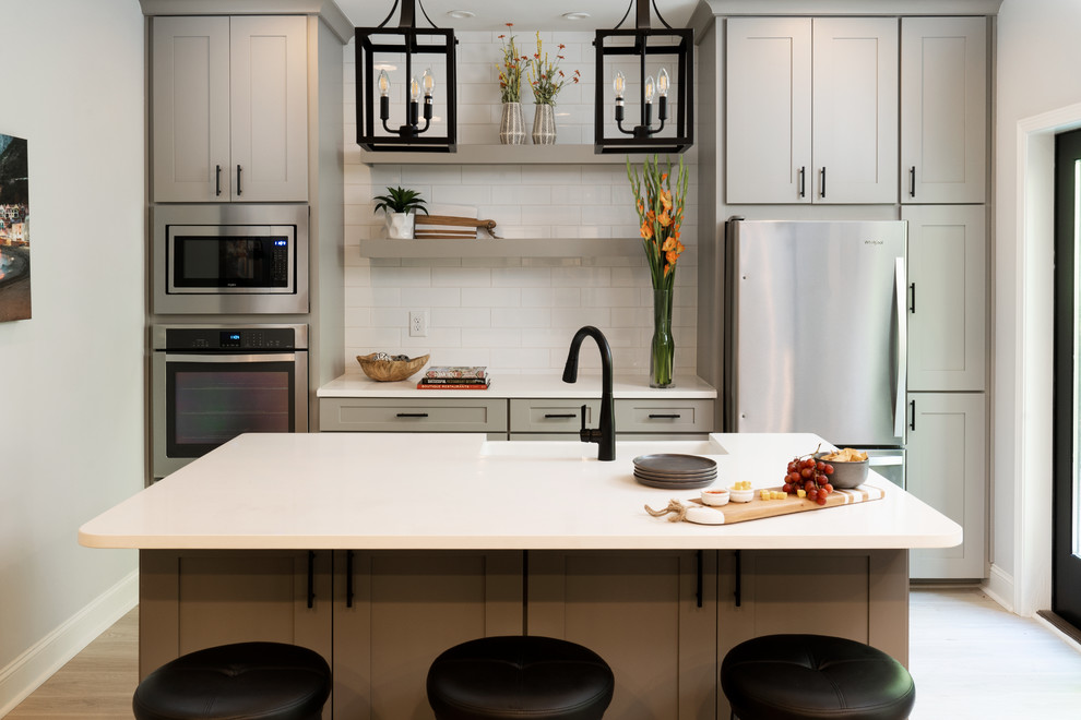 Example of a large transitional single-wall light wood floor and yellow floor open concept kitchen design with a farmhouse sink, shaker cabinets, gray cabinets, quartz countertops, white backsplash, ceramic backsplash, stainless steel appliances, an island and white countertops
