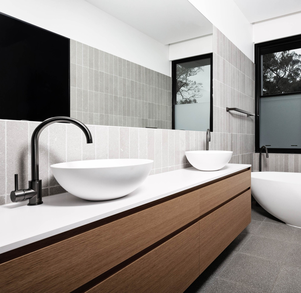 Photo of a medium sized modern ensuite bathroom in Adelaide with recessed-panel cabinets, medium wood cabinets, a freestanding bath, a one-piece toilet, grey tiles, metro tiles, white walls, ceramic flooring, a vessel sink, solid surface worktops, grey floors, white worktops, double sinks and a built in vanity unit.