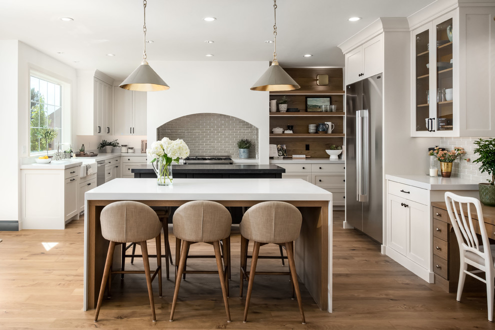Country eat-in kitchen in Salt Lake City with a farmhouse sink, shaker cabinets, white cabinets, grey splashback, subway tile splashback, stainless steel appliances, medium hardwood floors, multiple islands, brown floor and white benchtop.